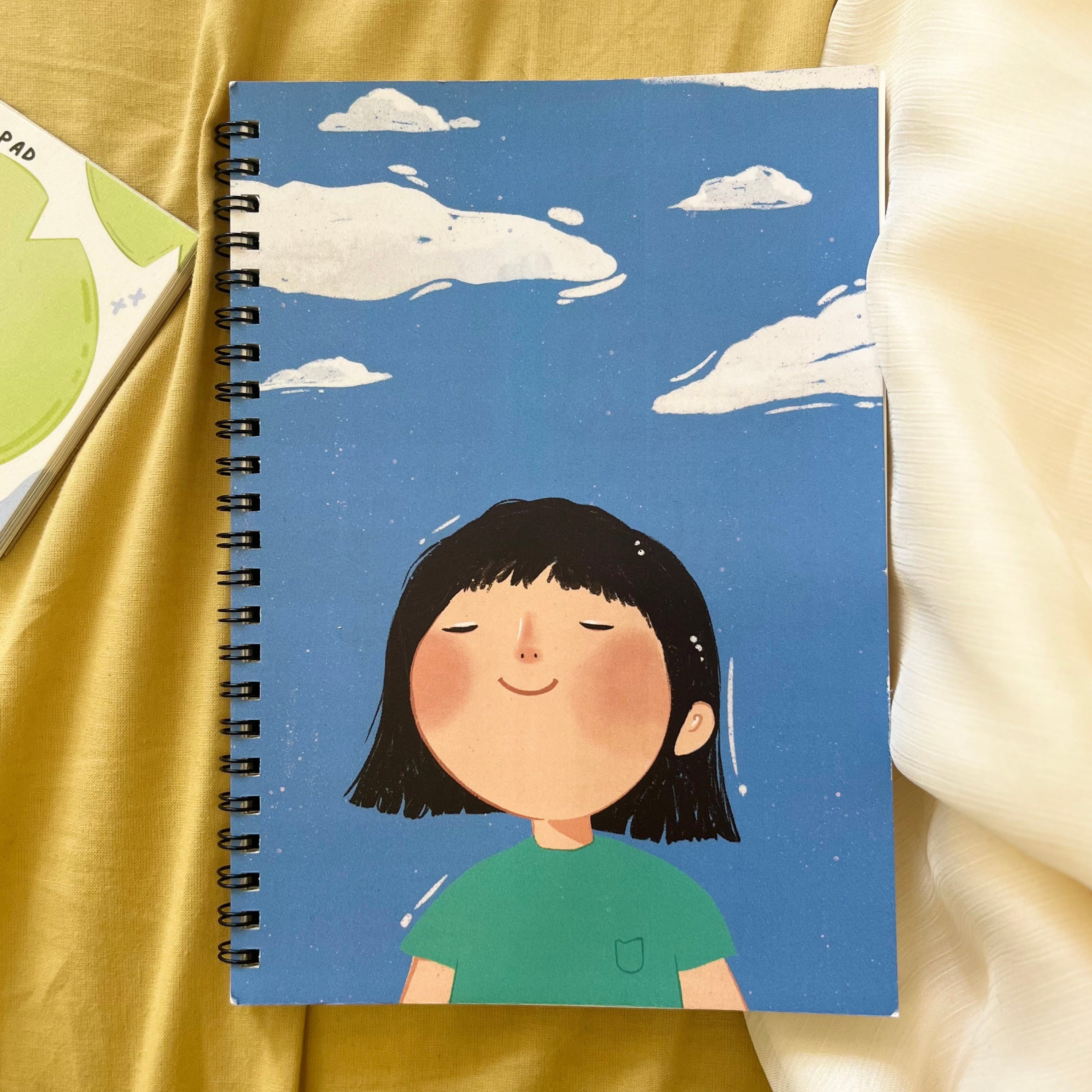 Notebook with a cartoon character on a blue background, placed on a yellow fabric surface.